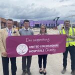 Four people are standing side-by-side in a line, holding a charity banner up. In the background is a building site with a partially built building and other building equipment.