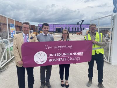 Four people are standing side-by-side in a line, holding a charity banner up. In the background is a building site with a partially built building and other building equipment. 