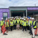 A group of people dressed in white hard hats and yellow high vis jackets stand in front of the new Lincoln Community Diagnostic Centre building. The building is still being constructed and is covered in a purple film.