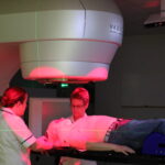 A patient lays on a clinical couch with a red light from a machine on them, with two staff stood by their head.