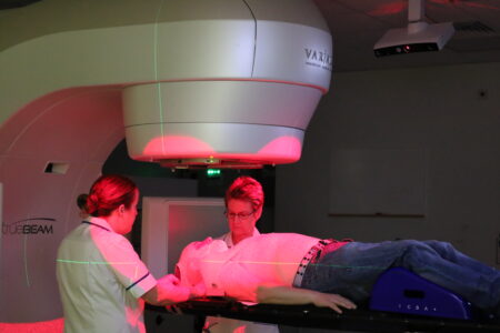 A patient lays on a clinical couch with a red light from a machine on them, with two staff stood by their head. 