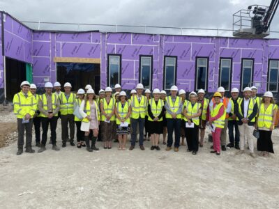 A group of people dressed in white hard hats and yellow high vis jackets stand in front of the new Skegness Community Diagnostic Centre building. The building is still being constructed and is covered in a purple film.