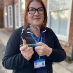 A woman with red hair and black glasses, wearing a nursing uniform, holds up her glass award trophy.