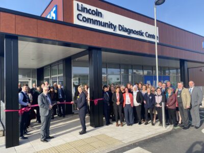 A group of people standing in from of a building. The Right Worshipful the Mayor of Lincoln, Councillor Alan Briggs, cutting the ribbon for Lincoln Community Diagnostic Centre with Professor Karen Dunderdale, Chief Executive for Lincolnshire Community and Hospitals Group.